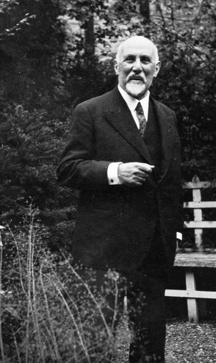 An elderly gentleman with white moutstache and beard, wearing a 3 piece suit with shrit and tie, standing in a garden in front of a wooden bench.
